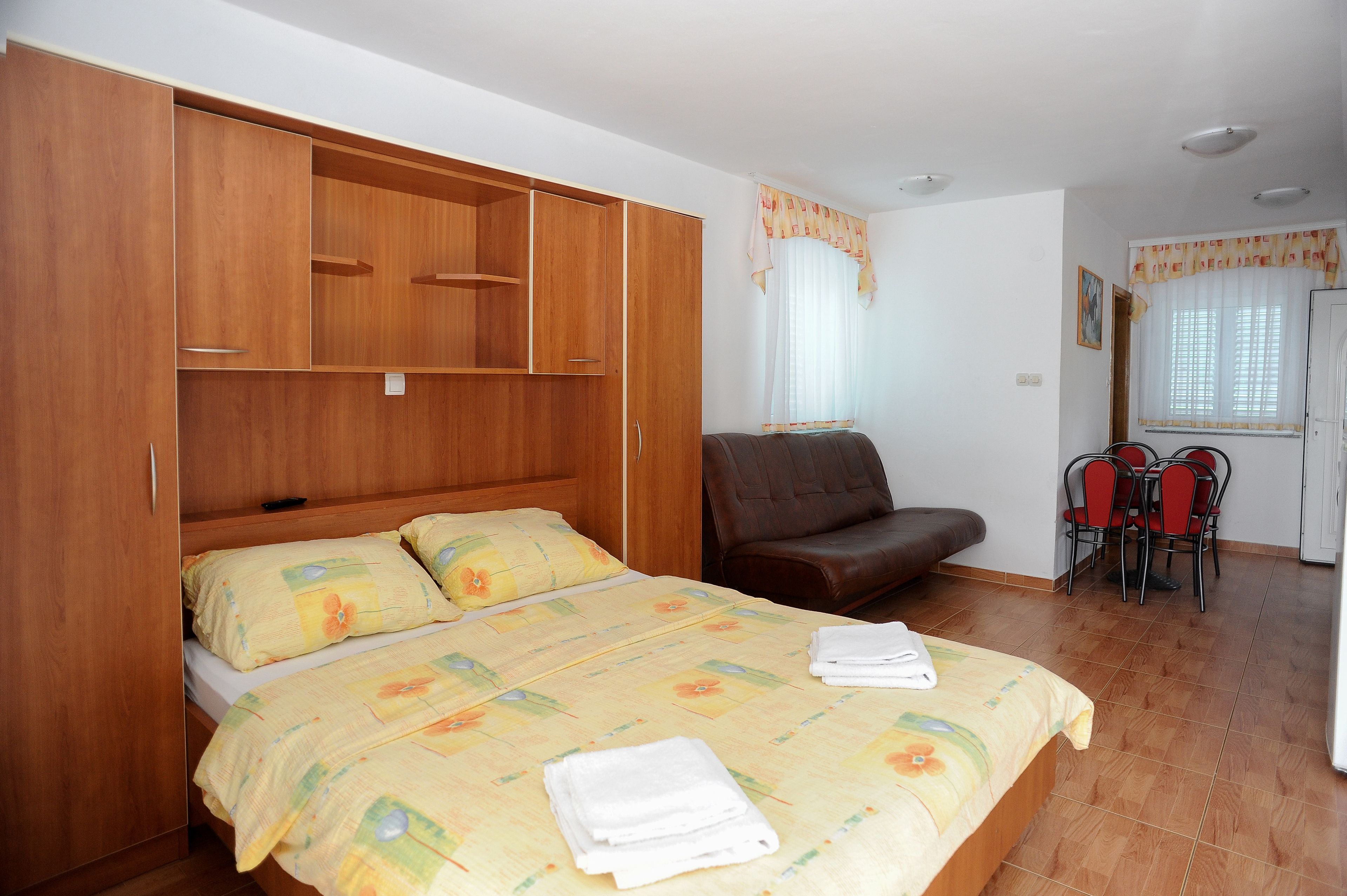 A bedroom with a bed, wooden wardrobe, a leather chair, a small dining table with chairs, and a window with curtains.