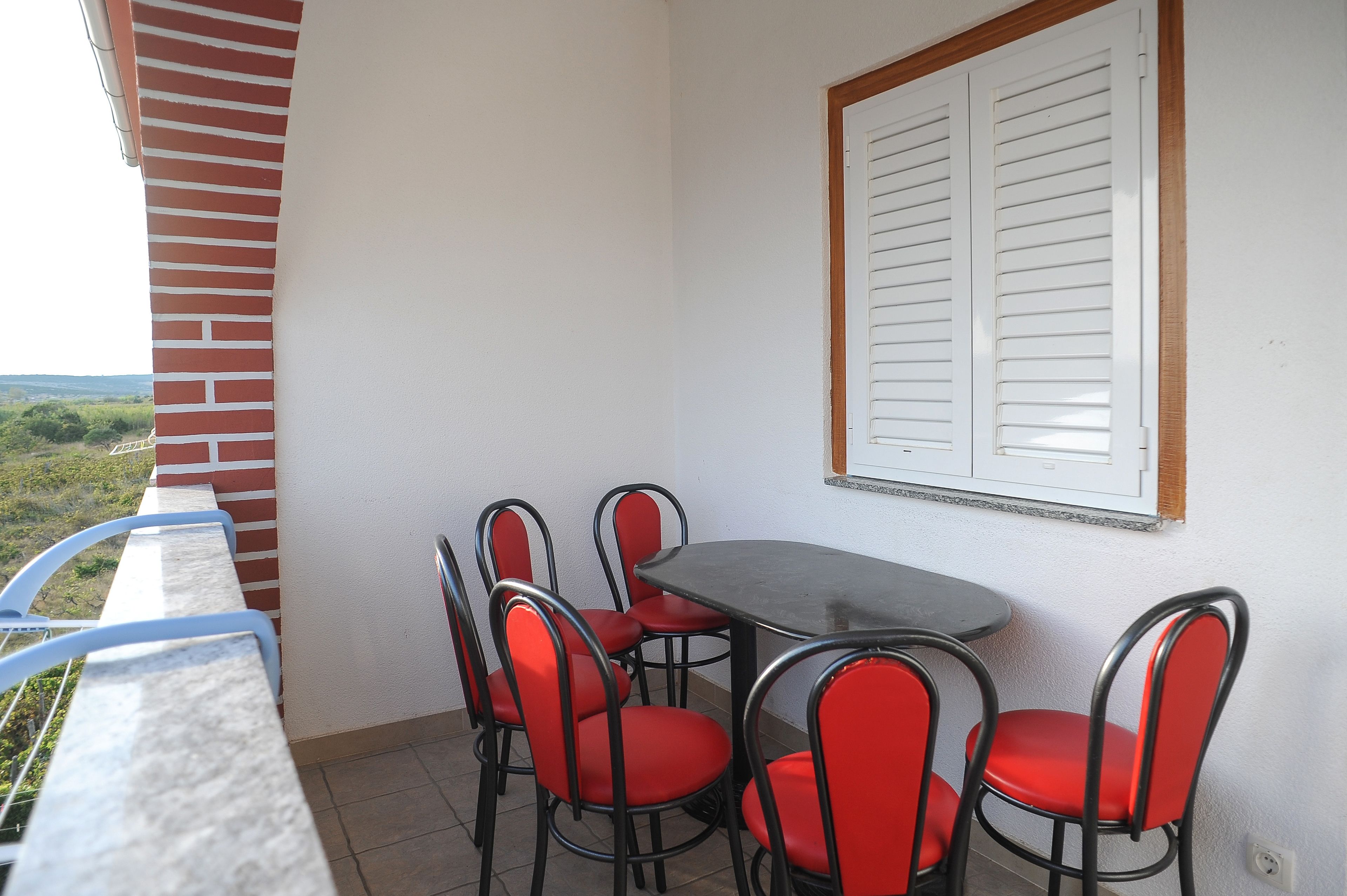 Un balcón con una mesa y cuatro sillas rojas, un muro blanco y una ventana con persianas.