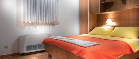 Un lit bien fait, avec une literie aux couleurs orange et jaune, une armoire en bois et une fenêtre ornée de rideaux légers.