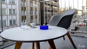 A round table with a white top and wooden legs, a gray chair, a blue ashtray, and a glass of water on a balcony with a view of buildings and a street. - BlueStone Boarding Apartments (Düsseldorf)