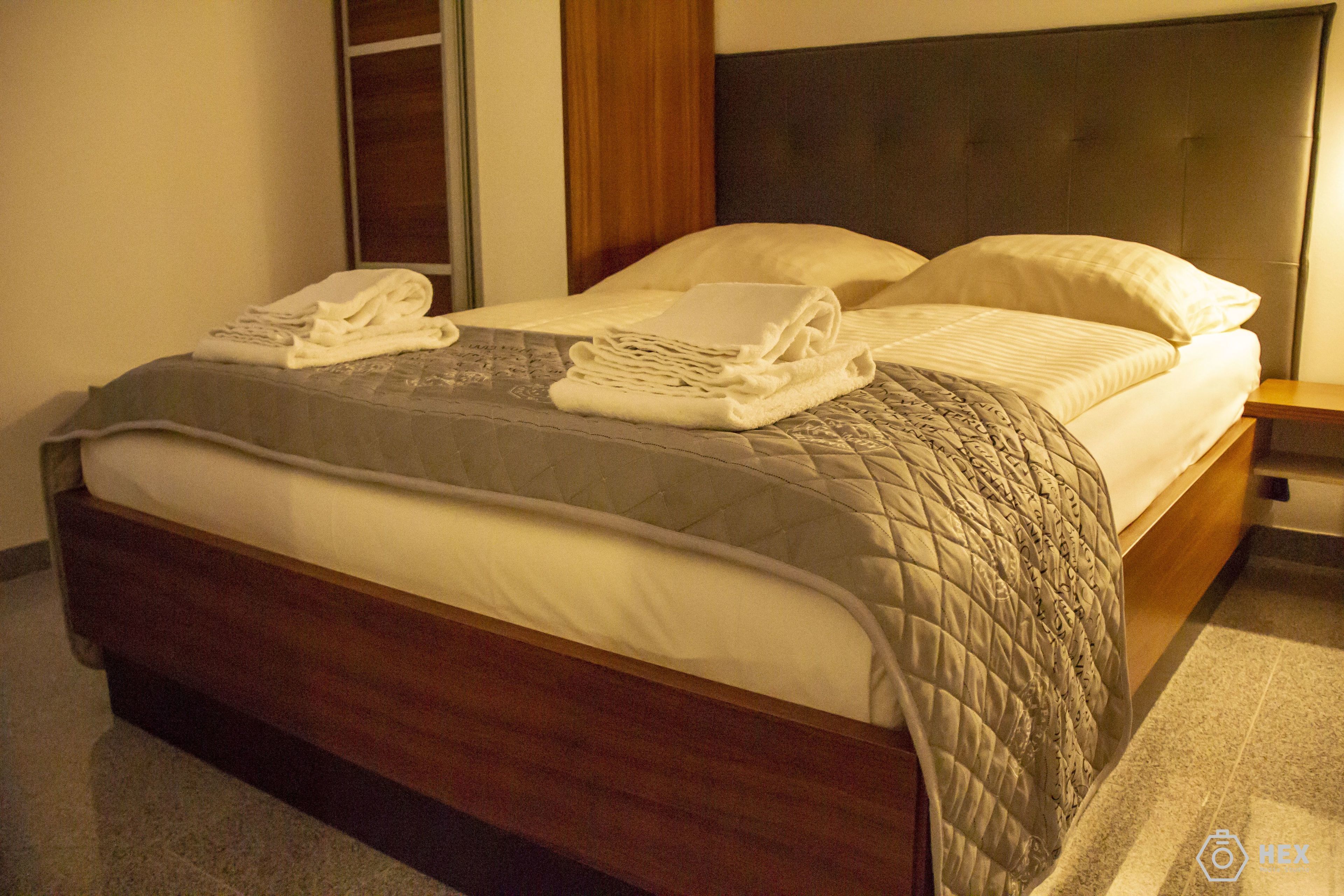 Ein ordentlich bezogenes Bett mit gesteppter Decke und gefalteten Handtüchern, ein Holz-Kopfteil und ein Nachttisch mit Lampe.