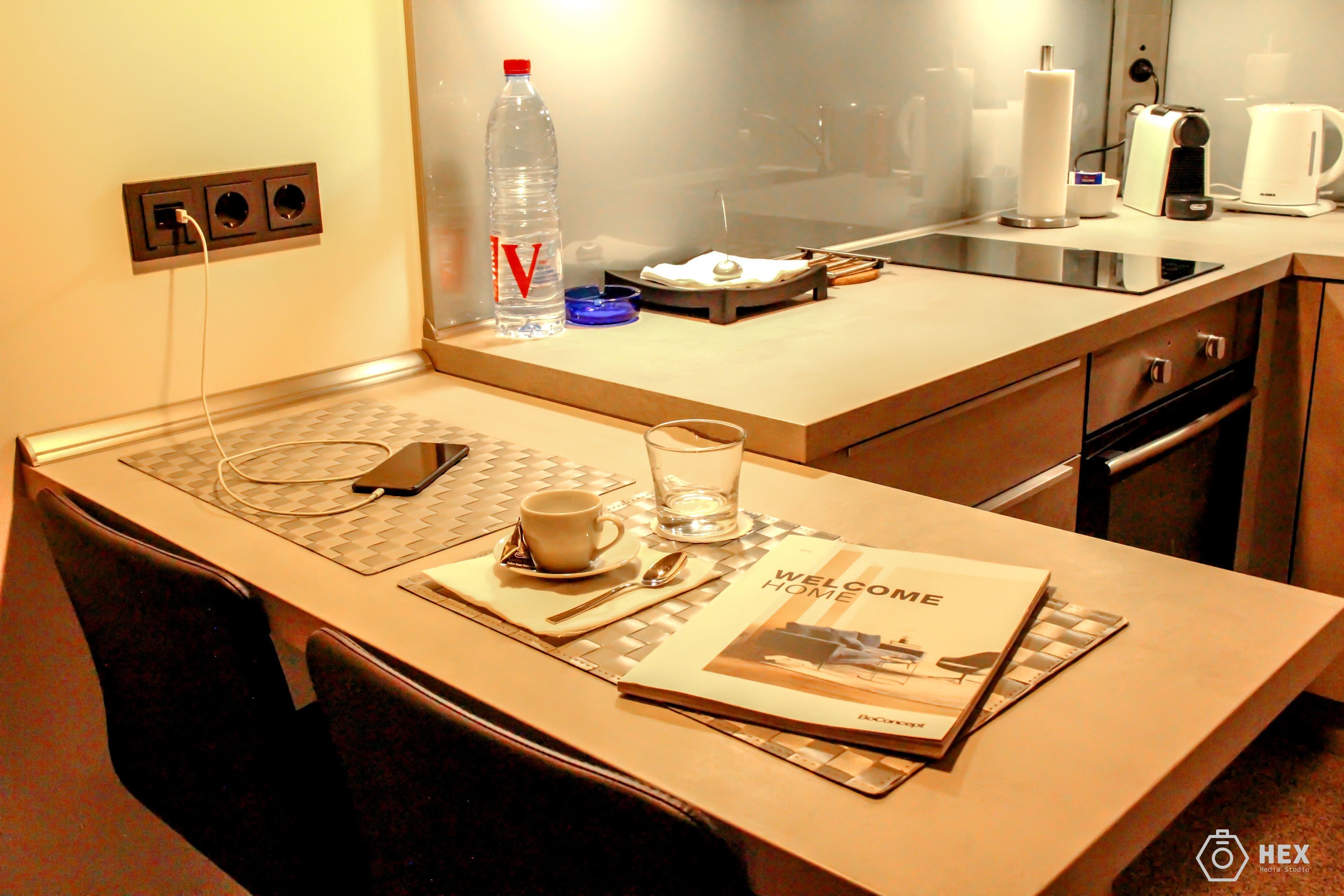 A hotel room with a small kitchenette, a welcome home booklet, a bottle of water, a glass, and a cup on the counter.
