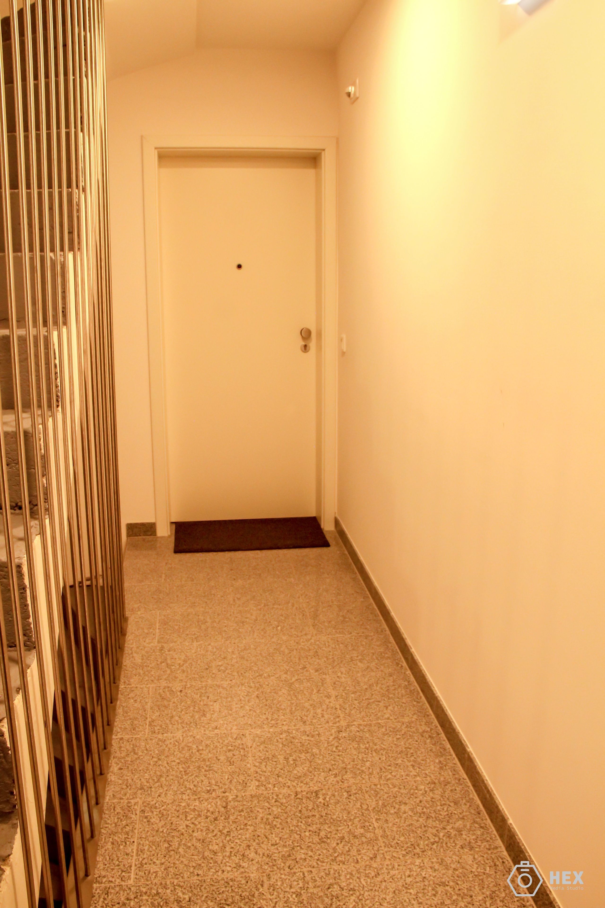 Um corredor estreito com uma porta branca no final, um corrimão de metal à esquerda e piso coberto de carpete.