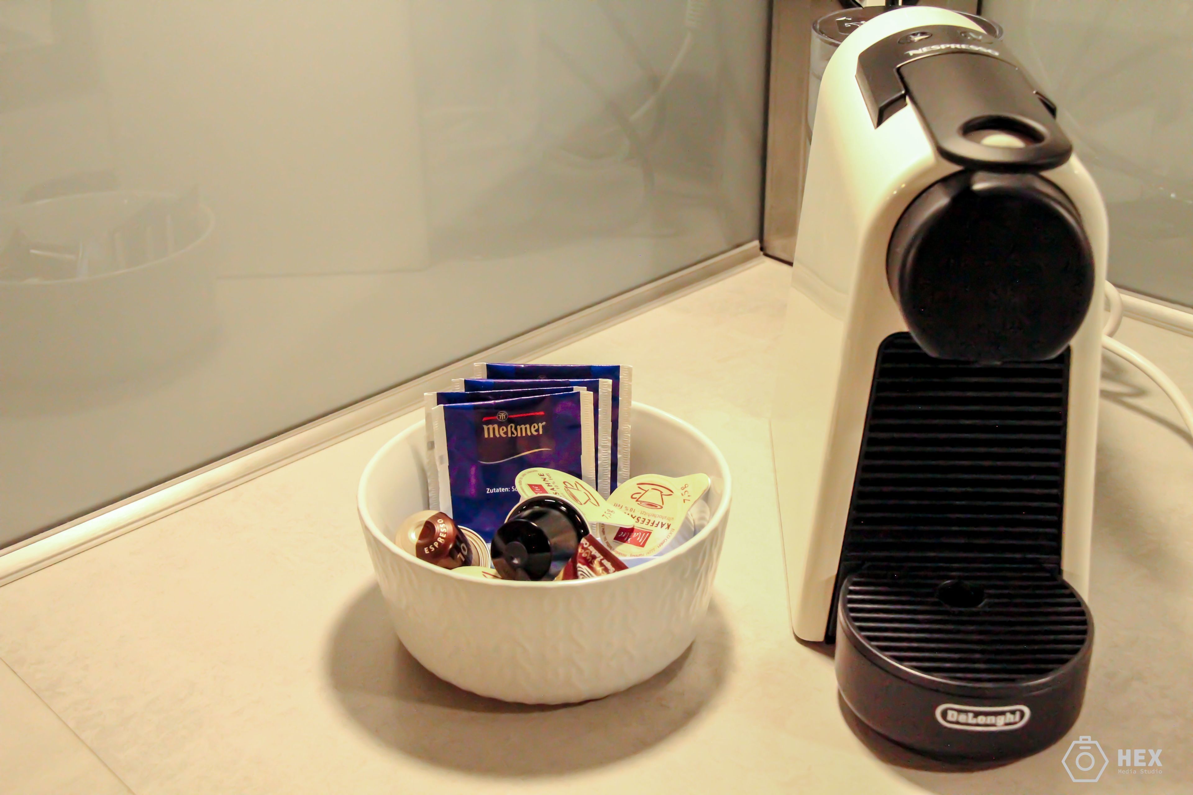 A white bowl containing various items including a bottle, a pack of Meijer chocolate, and some cards, placed next to a black and white coffee maker on a tiled surface.
