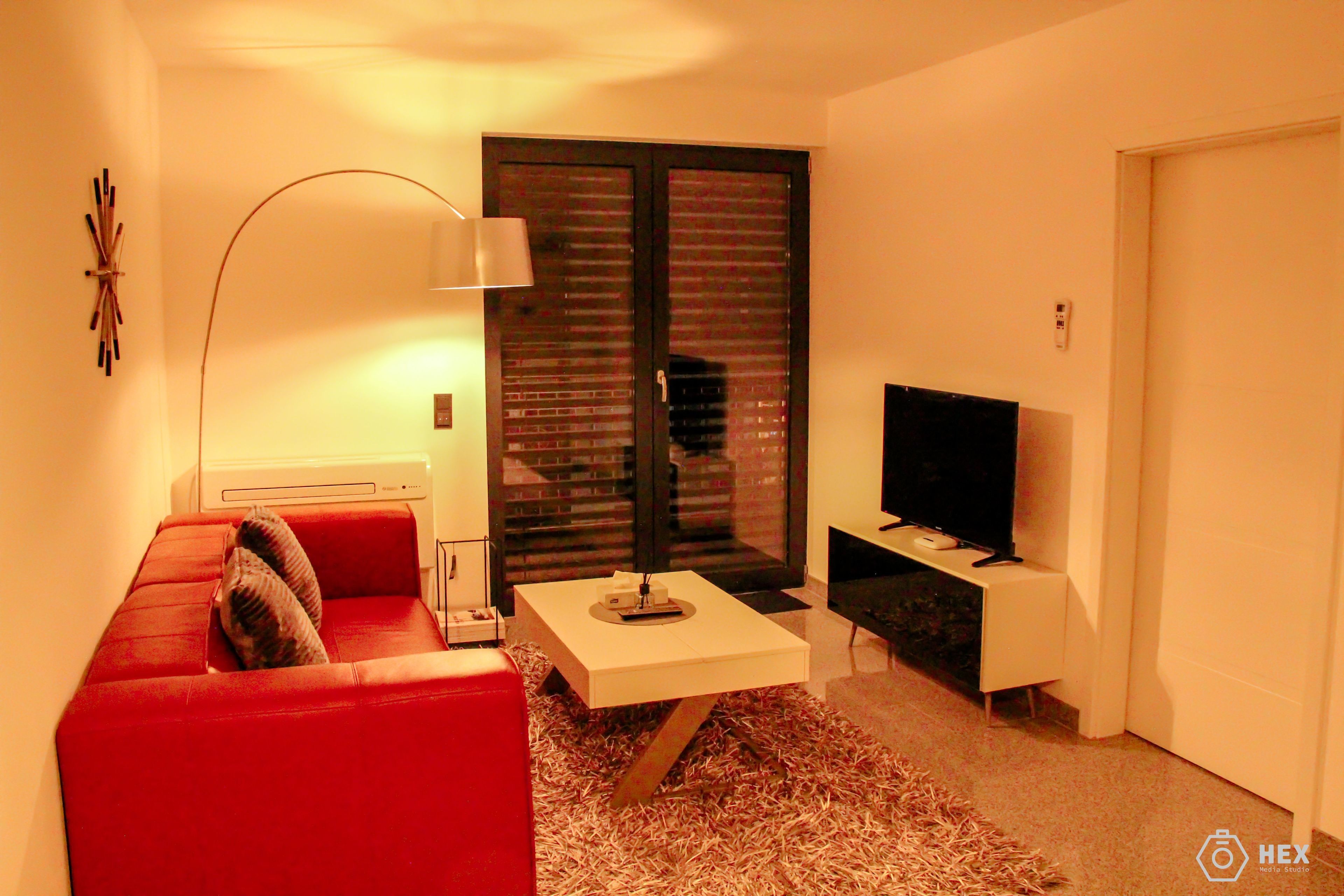 A modern living room with a red sofa, a coffee table, a TV, and a large window with blinds.