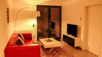 A modern living room with a red sofa, a coffee table, a TV, and a large window with blinds.