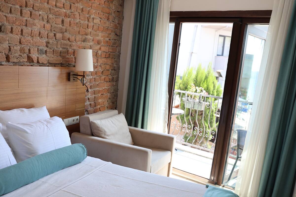 A hotel room with a large bed, a chair, a table, and a balcony with a view.