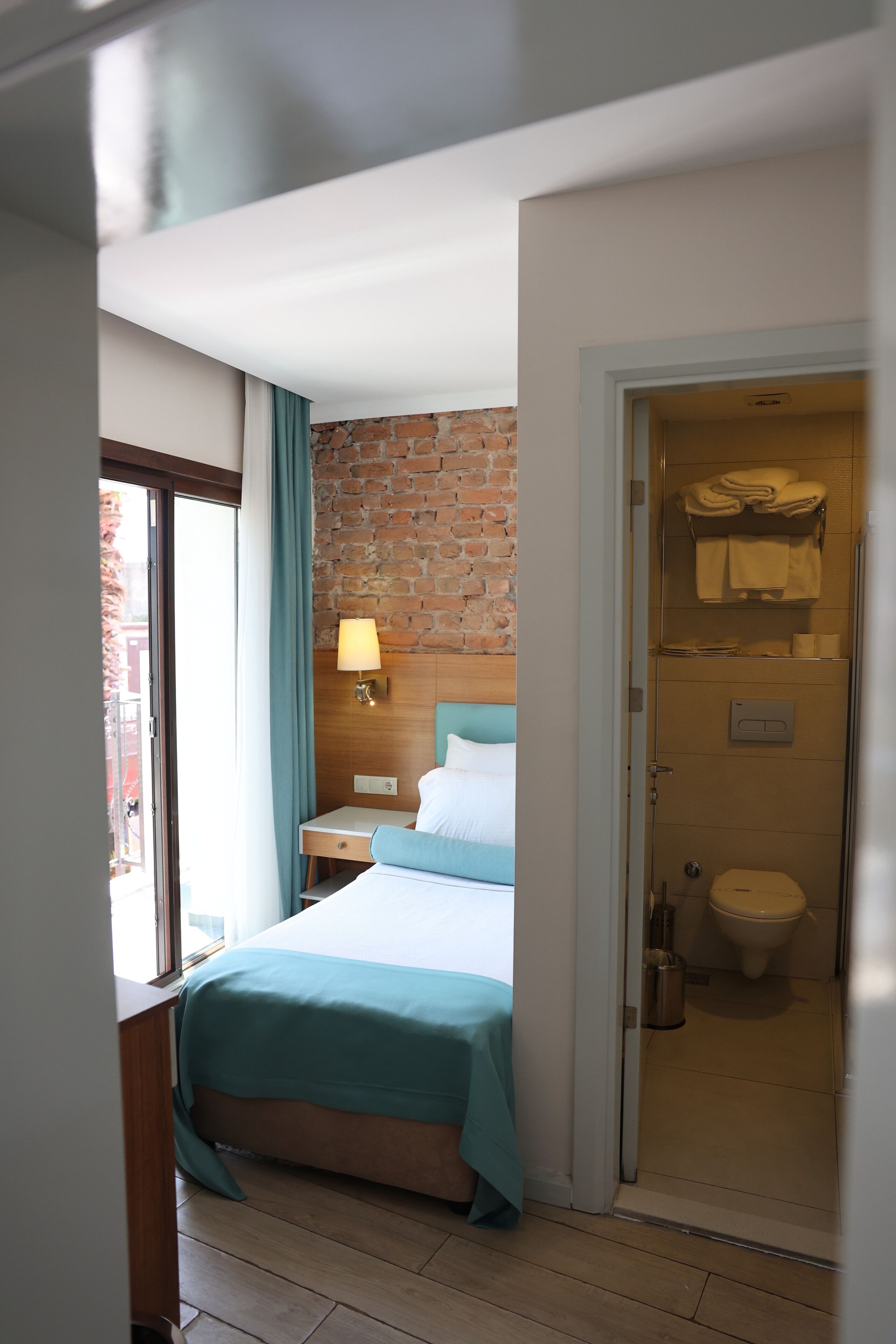 A hotel room with a bed, a nightstand, a lamp, a brick wall, and a bathroom with a toilet and towel rack.