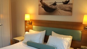 Habitación de hotel con una cama grande, dos mesitas de noche, un cabecero de madera y un cuadro de un barco en la playa.