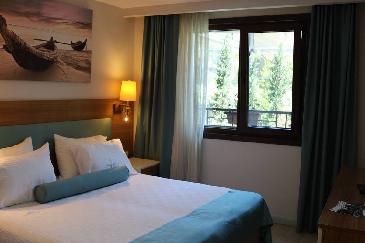 A hotel room with a large bed, a nightstand with a lamp, a window with curtains, and a painting on the wall.