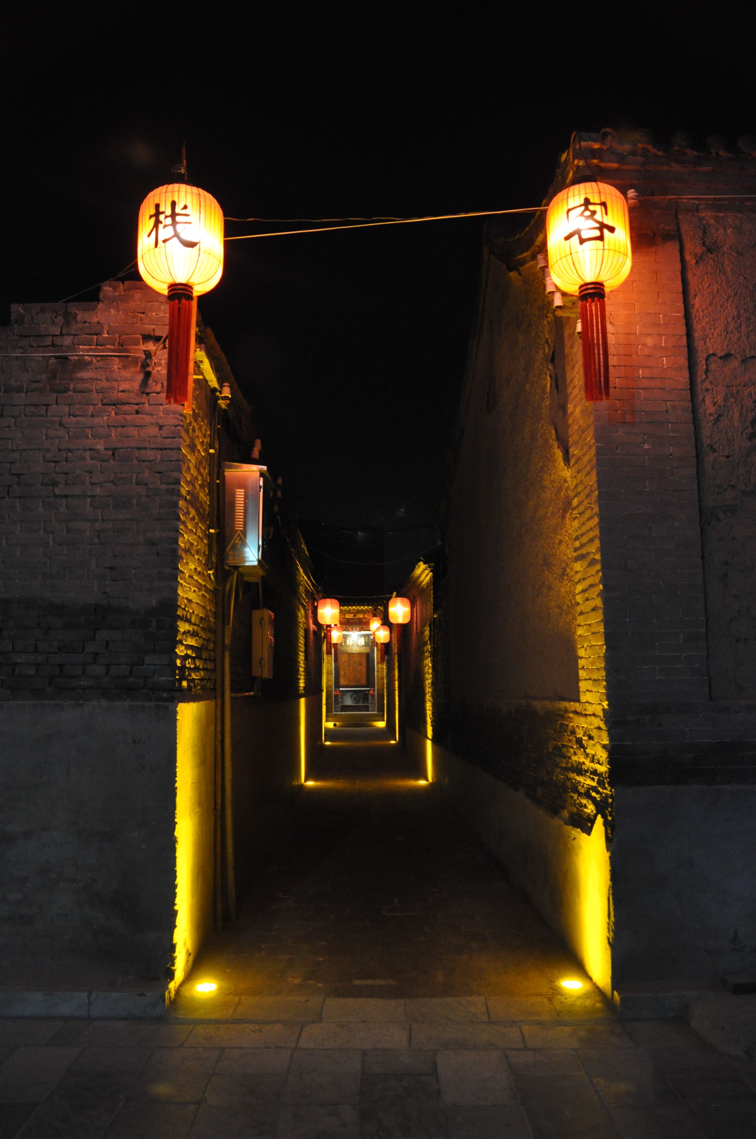 Une ruelle étroite, bordée de murs de briques, où des lanternes traditionnelles sont suspendues au plafond.