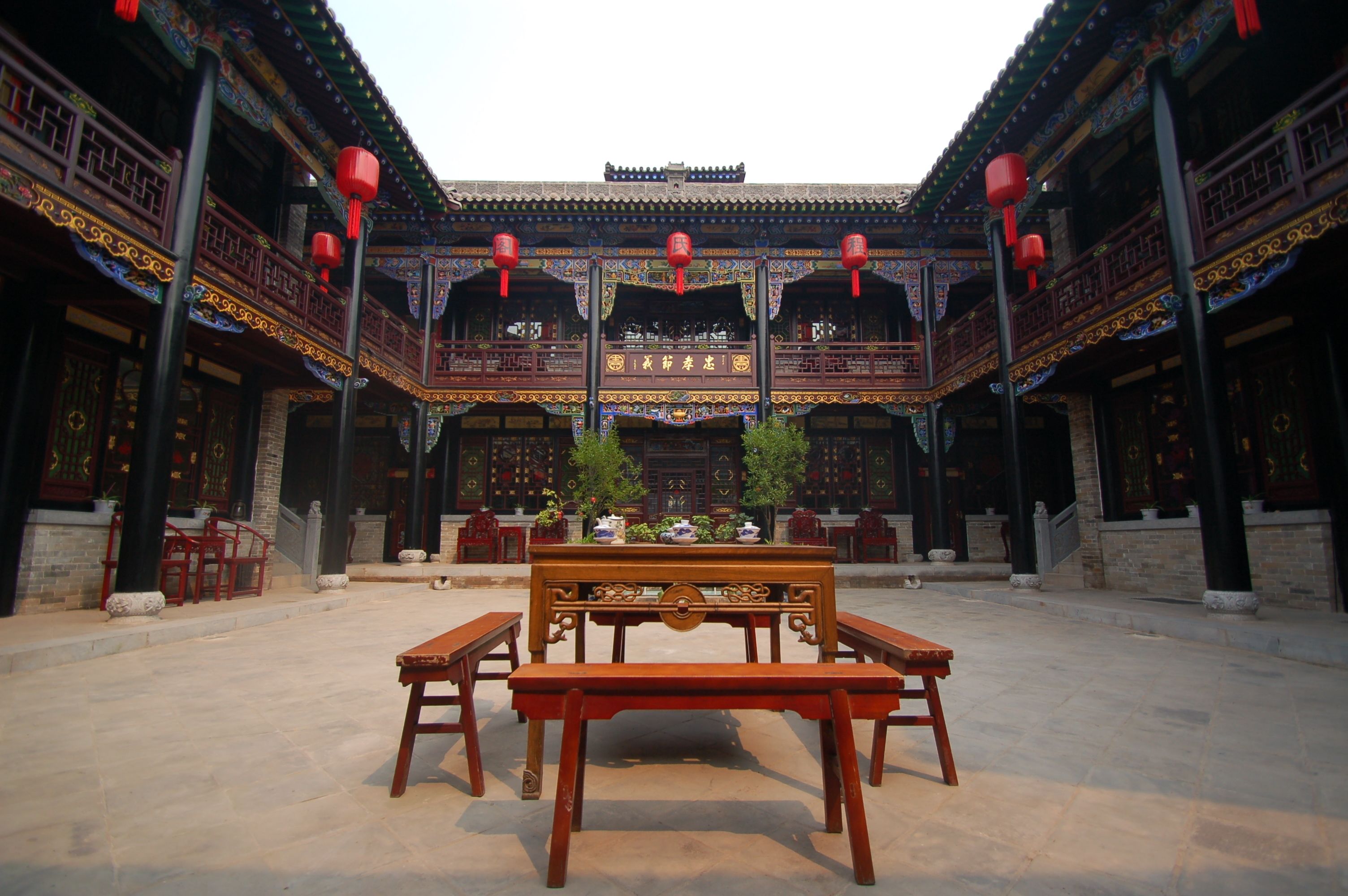 Une cour traditionnelle avec une table centrale et des bancs, entourée de bâtiments richement décorés et illuminés par des lanternes rouges.