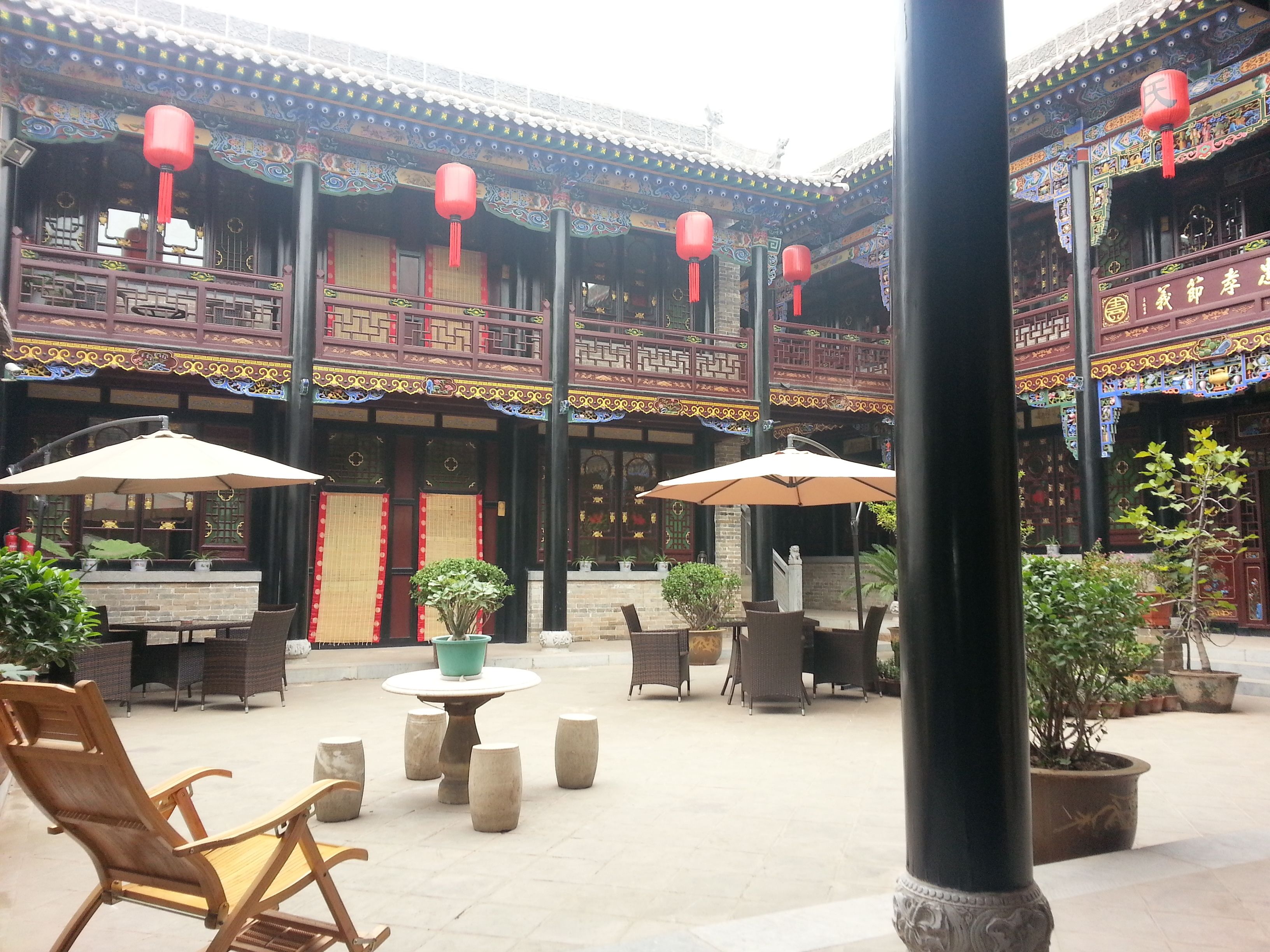 An outdoor courtyard with traditional Chinese architecture, featuring ornate wooden balconies, red lanterns, and potted plants.