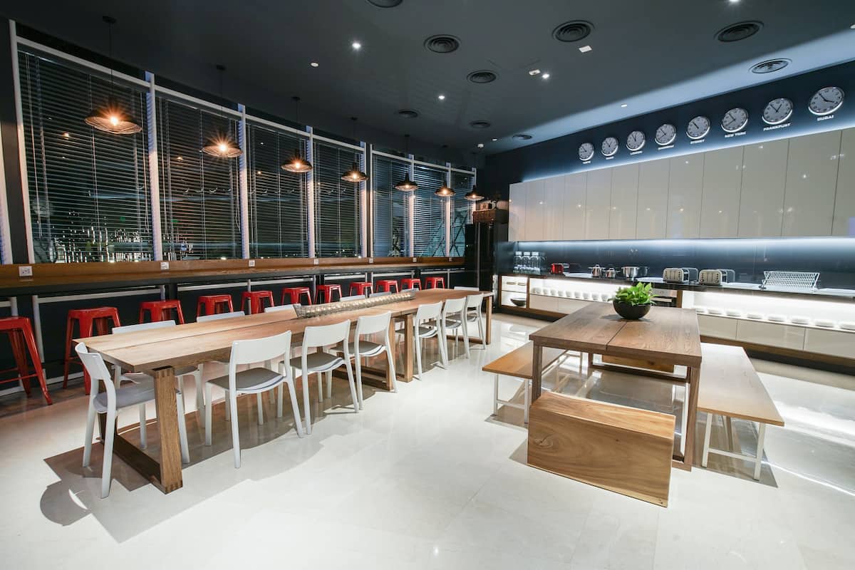 Un restaurante moderno con una larga mesa de madera y sillas rojas, ventanas amplias con persianas y una pared con relojes.