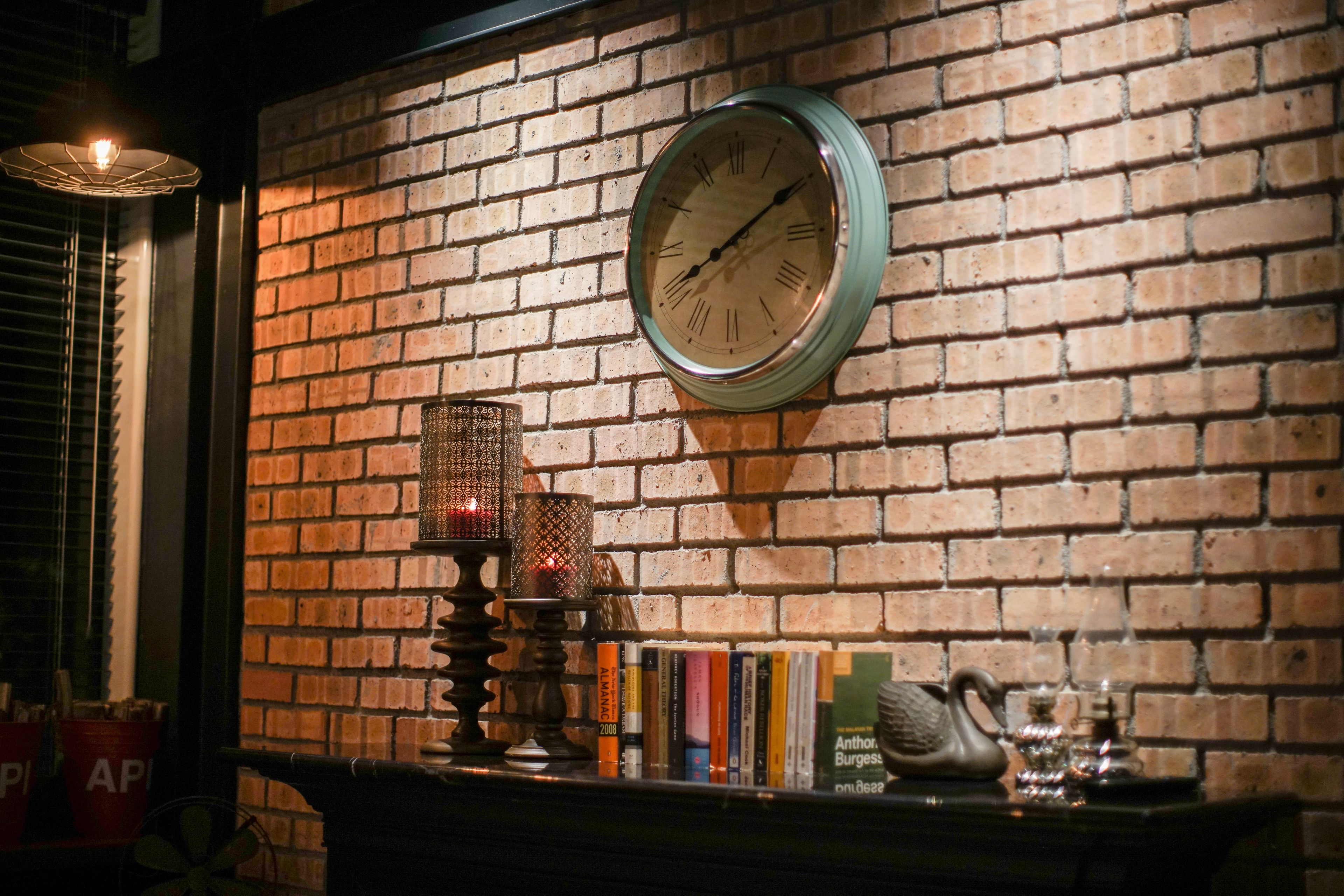 A brick wall with a round clock, a bookshelf with books, and a decorative candle holder.