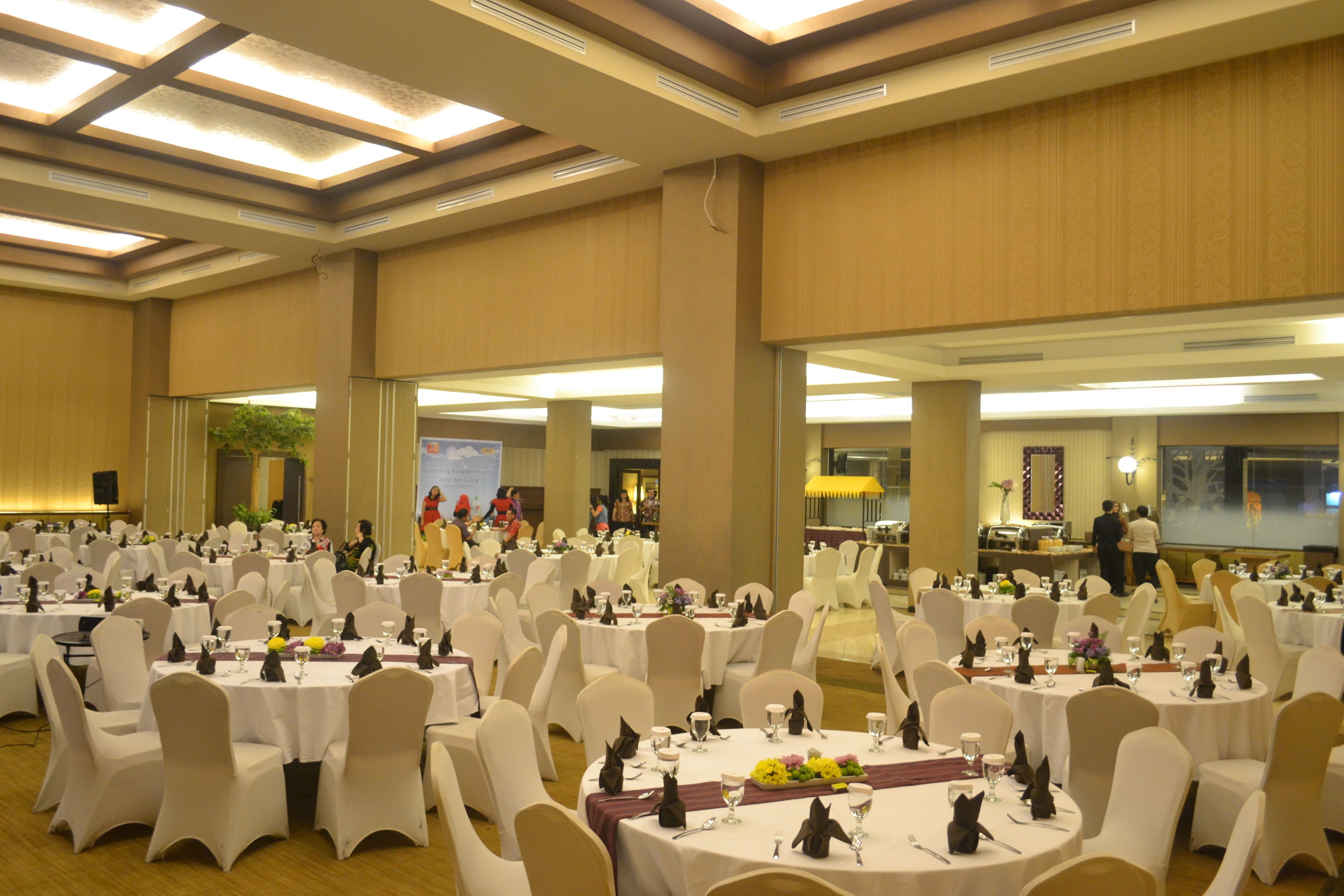 A large banquet hall with round tables and white chairs, equipped with table settings and a stage in the background.