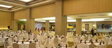A large banquet hall with round tables and white chairs, equipped with table settings and a stage in the background.