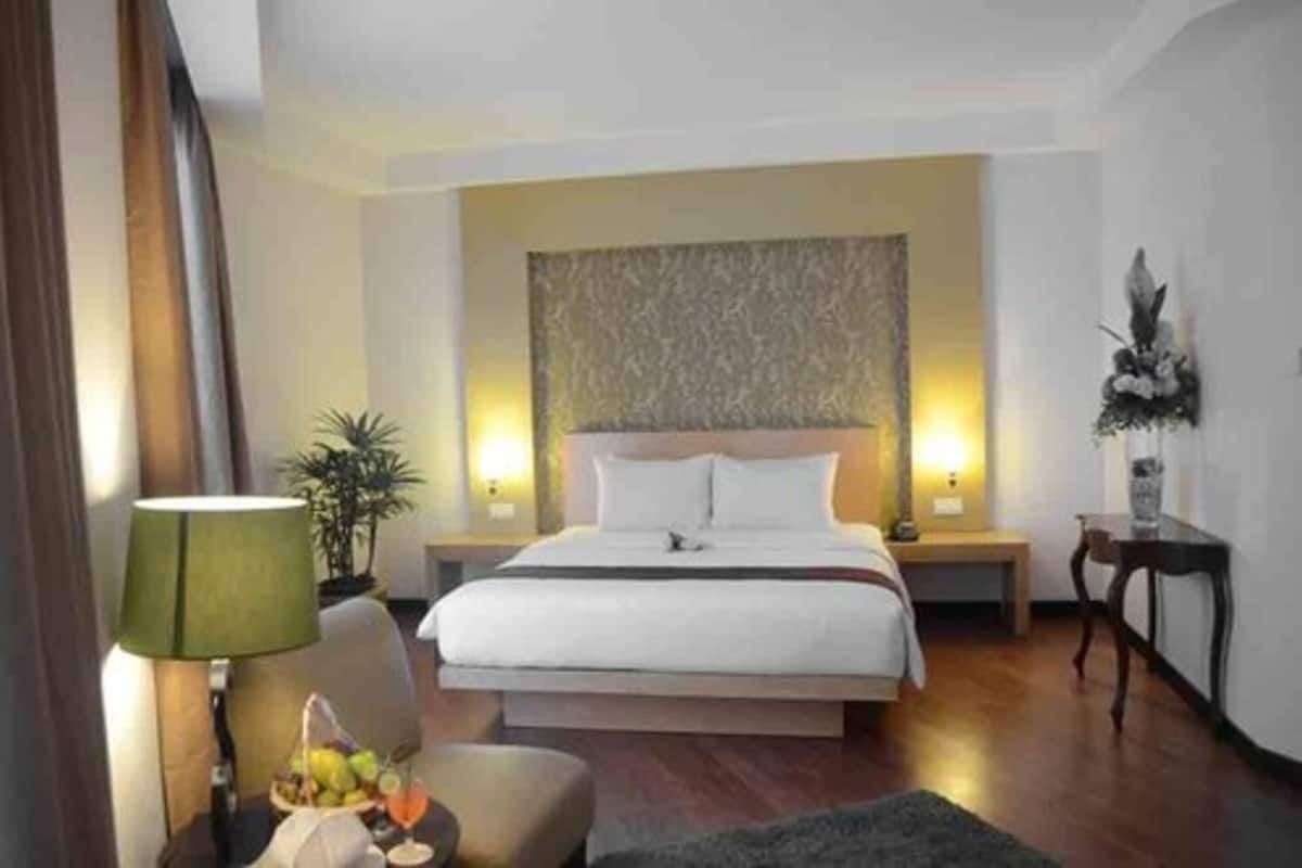 A hotel room with a large bed, bedside tables, a lamp, a plant, and a small table with a fruit basket and drink.