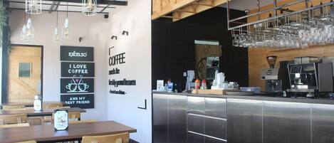 L'intérieur d'un café moderne, avec des tables et des chaises en bois, un comptoir en acier inoxydable et des luminaires suspendus.