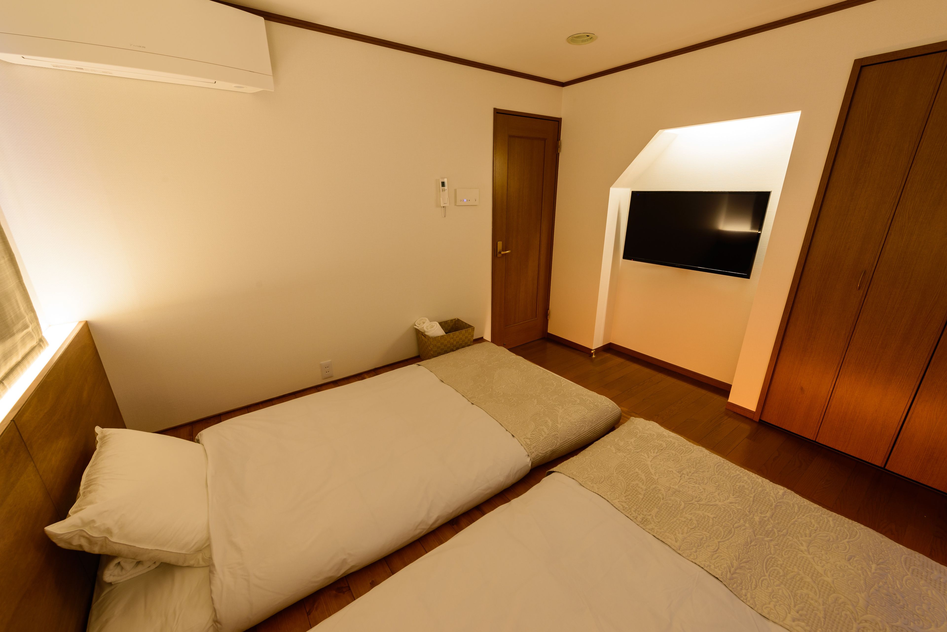 Habitación de hotel con dos camas, un televisor montado en la pared, un armario de madera y una mesita de noche pequeña.