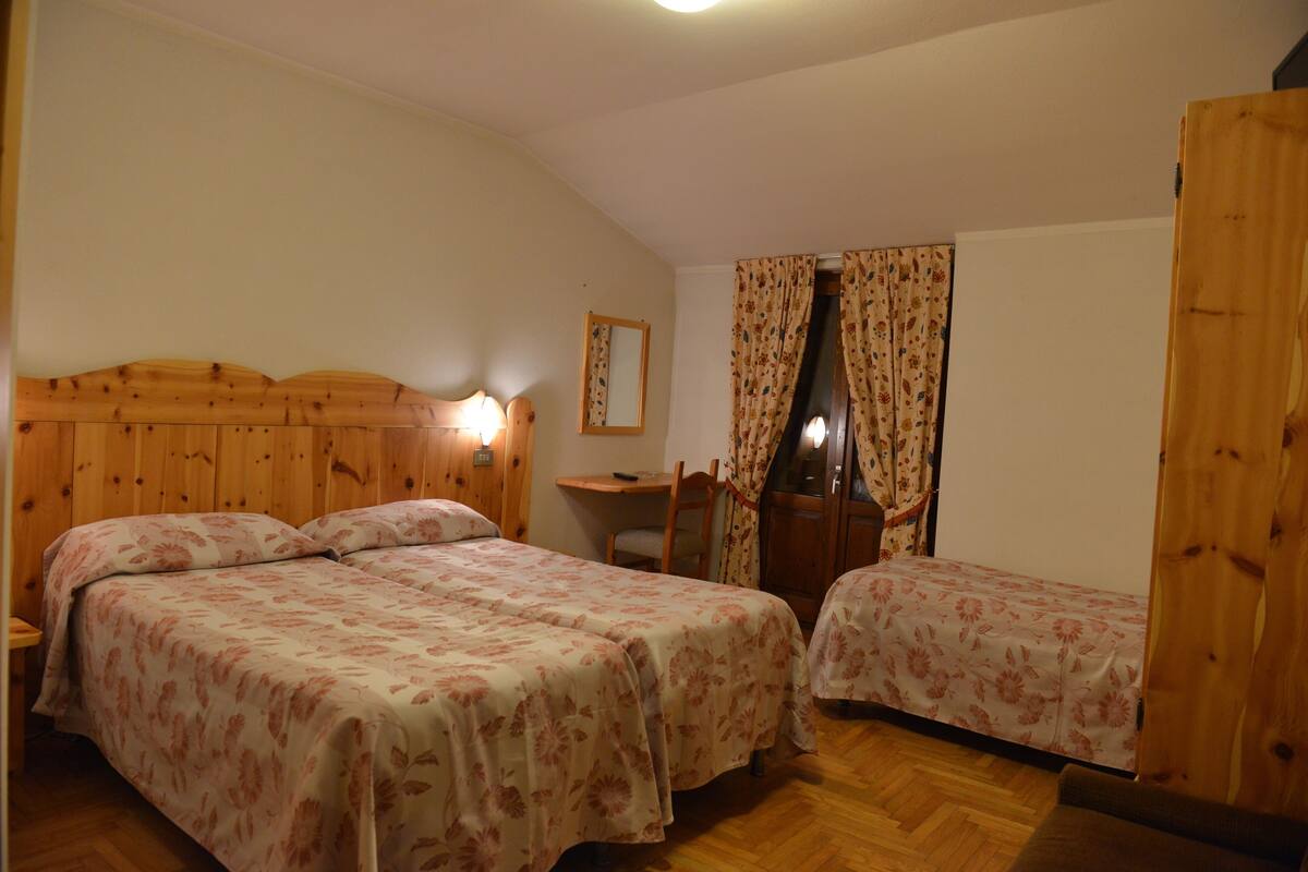 Ein Zimmer mit zwei Betten, einem Holzkopfende, einem Schreibtisch mit Stuhl, einem Spiegel und einem Fenster mit Vorhängen.