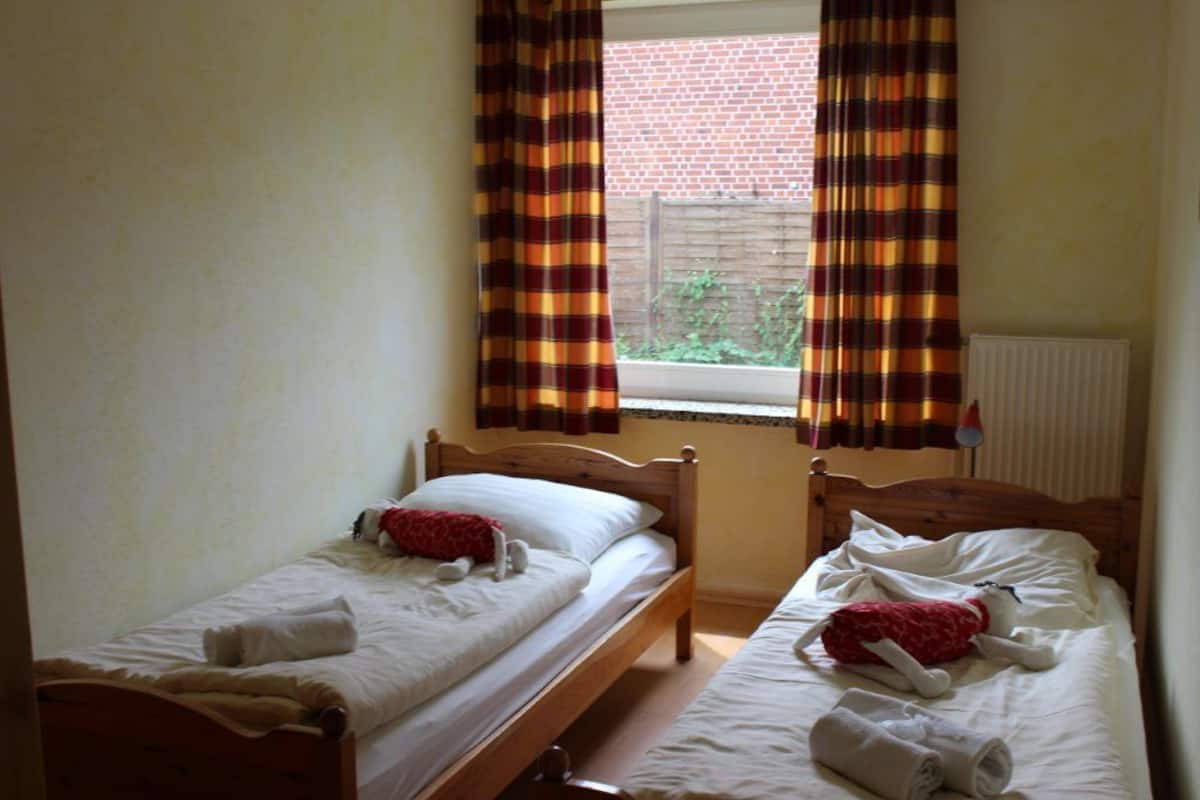 Un petit logement composé d’une seule pièce, avec deux lits simples, une fenêtre avec des rideaux et un radiateur.