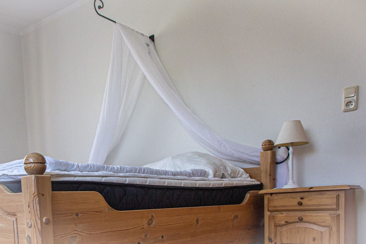 Un lit en bois surmonté d’un baldaquin blanc, une table de chevet avec une lampe et une prise électrique murale.
