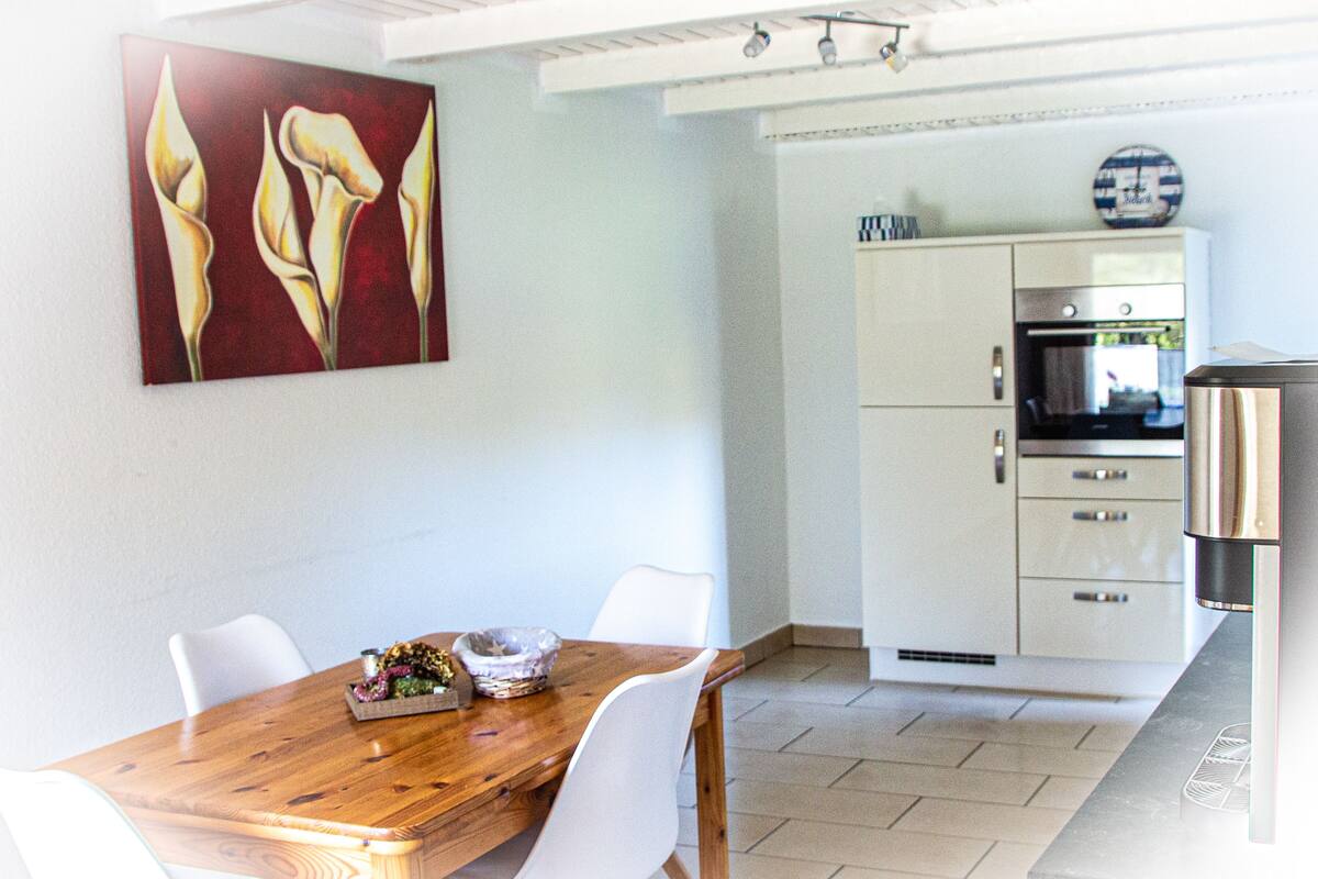 Un coin repas avec une table en bois et des chaises blanches, une cuisine avec des meubles blancs et des appareils en acier inoxydable, et un tableau représentant des lys calla, accroché au mur.
