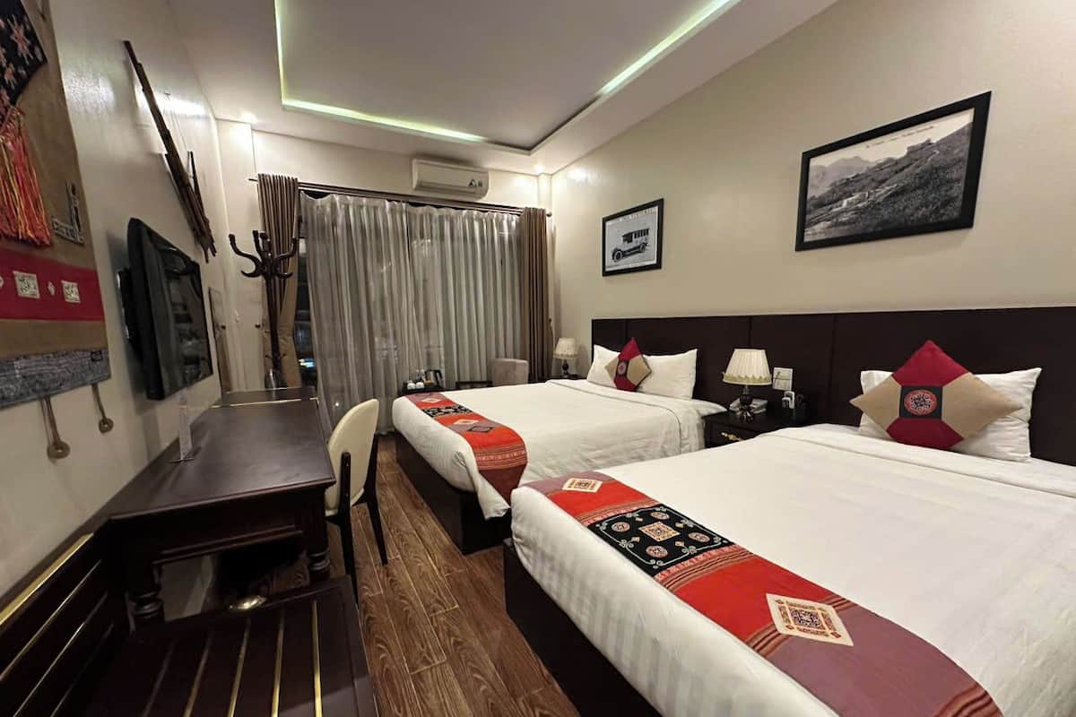 Habitación de hotel con dos camas, un escritorio y una silla. Cuenta con ventana con cortinas, aire acondicionado de pared y un cuadro enmarcado.
