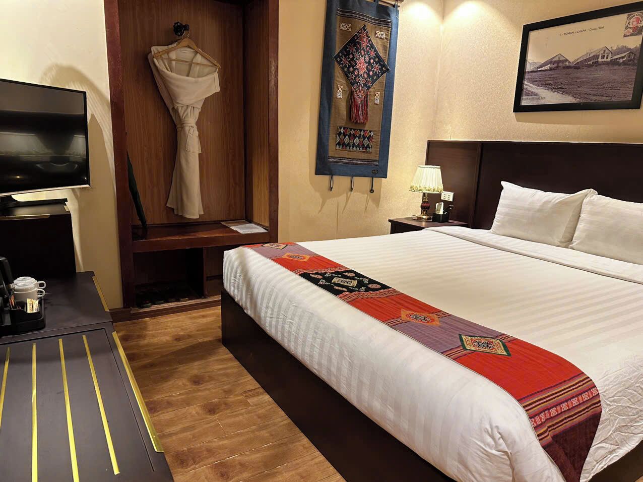 A hotel room with a large bed, a television on a desk, a coat rack, and a framed picture on the wall.