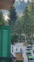 A street scene with a green building in the foreground, a busy road with vehicles and pedestrians, and trees in the background.