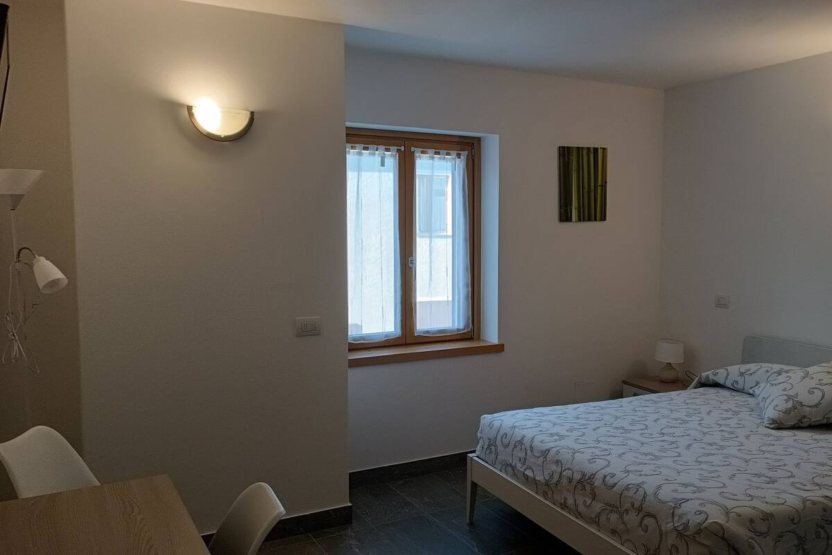 A bedroom with a bed, a nightstand, a chair, a window with curtains, and wall-mounted lights.