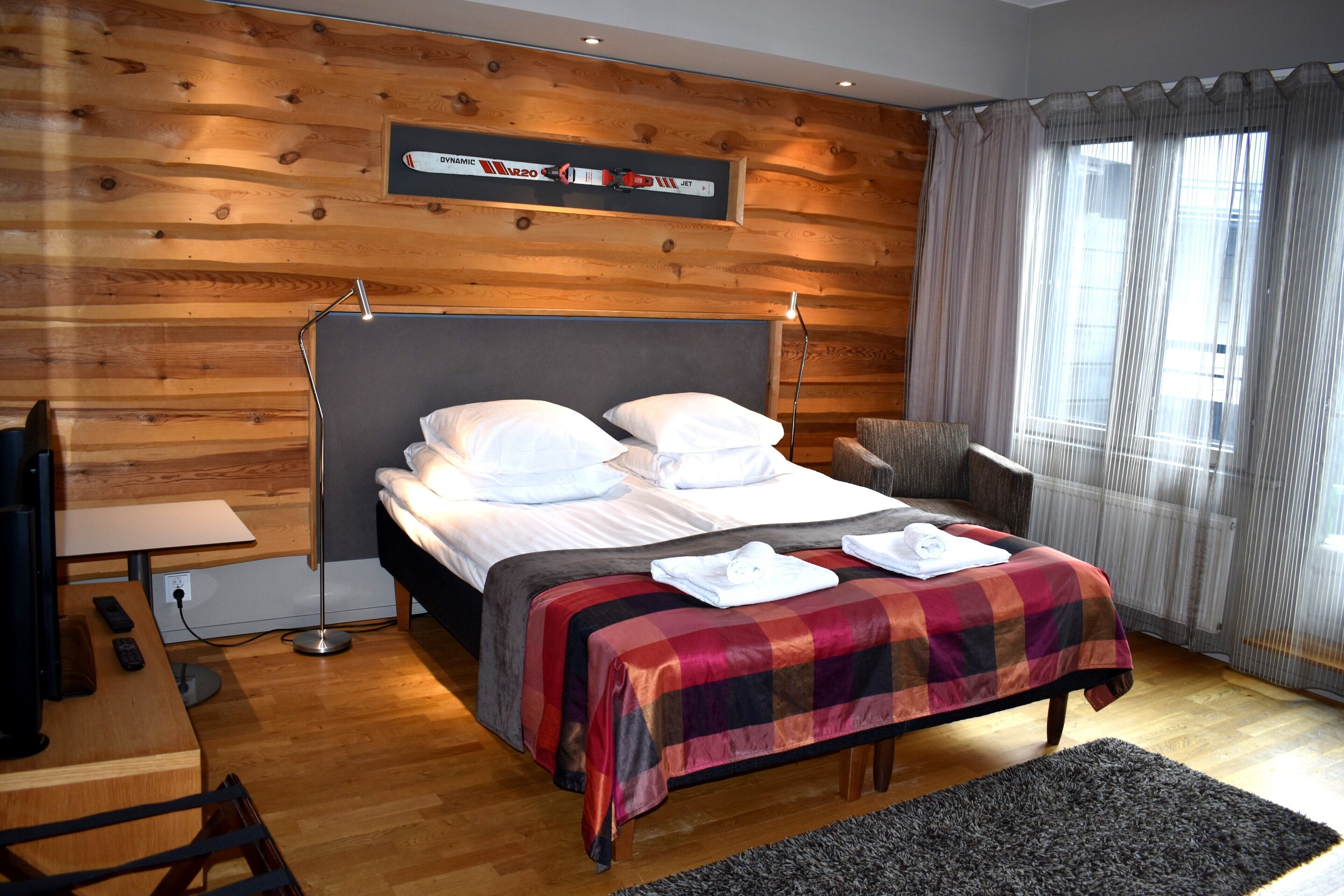 Een slaapkamer met een houten wandpaneel, een bed met een geruite spreideken, een nachtkastje met een lamp, en een raam met dunne gordijnen.