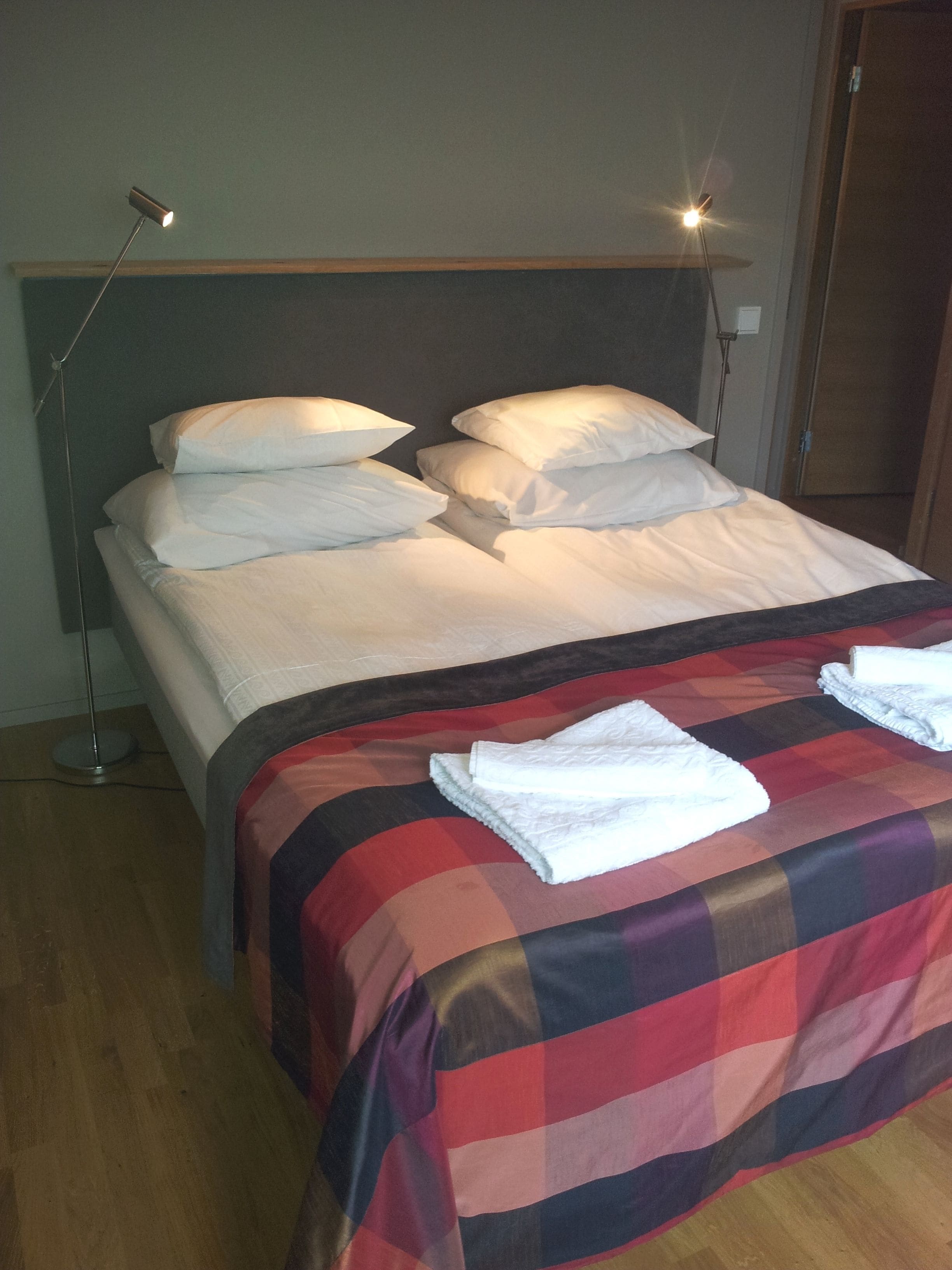Una cama con sábanas blancas y una manta escocesa, dos lámparas de noche y un suelo de madera.