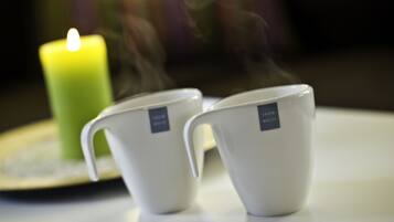 Two white ceramic cups with labels on a white surface, a lit green candle, and a blurred background.