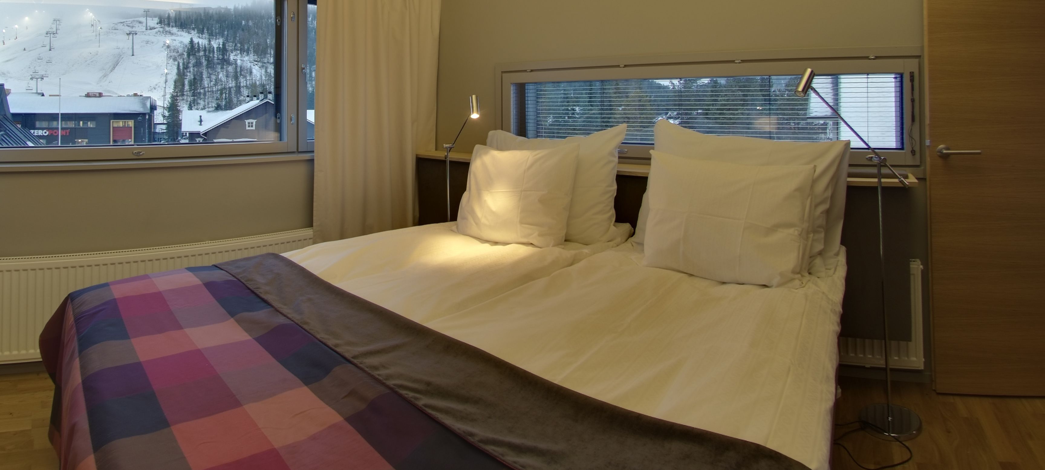 A hotel room with a bed, a window with blinds, a lamp, and a view of a snowy landscape.