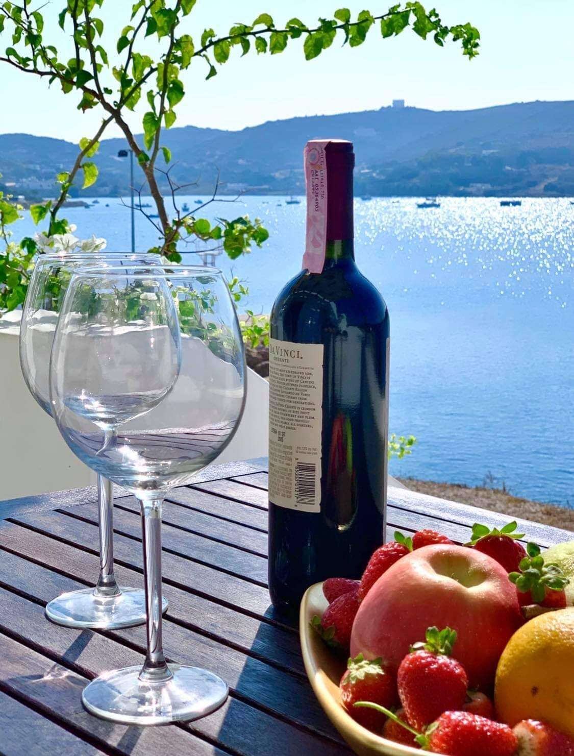 A table with two empty wine glasses, a bottle of wine, and a bowl of fruit by a lakeside view.