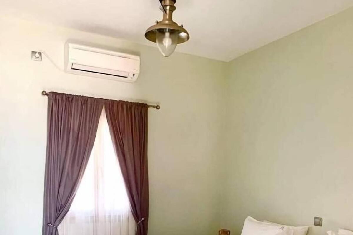 Une chambre d’hôtel avec un lit, du linge de lit blanc, un ventilateur de plafond, un appareil de climatisation et une fenêtre avec des rideaux.