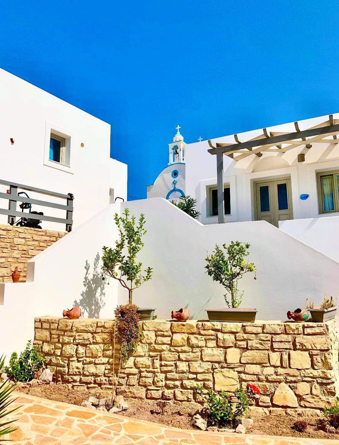 Un edificio blanco con muro de piedra, plantas en macetas y un cielo azul despejado.