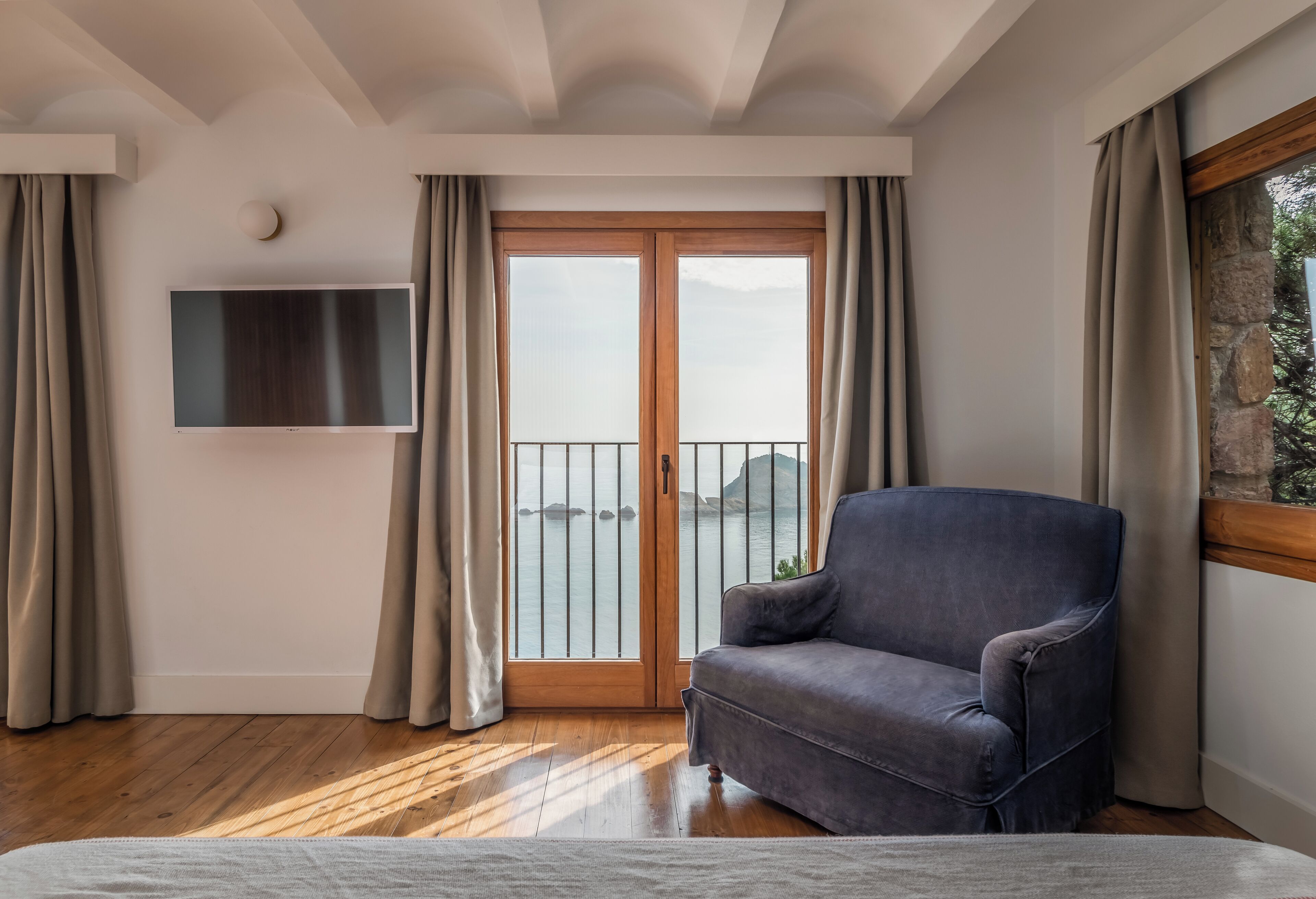 Una habitación con suelo de madera, un sofá gris, un televisor montado en la pared y un balcón con vistas al mar.