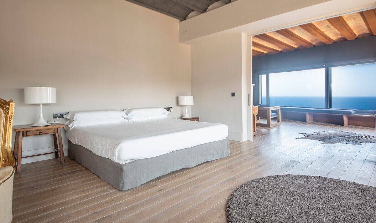 A bedroom with a large bed, wooden flooring, and a view of the ocean.
