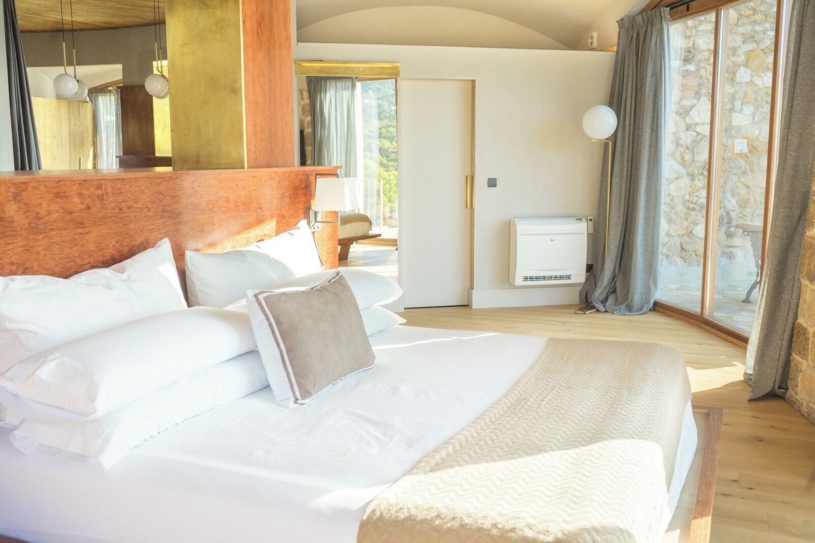 A large bed with white linens and a brown pillow, a wooden headboard, and a stone wall with a window.