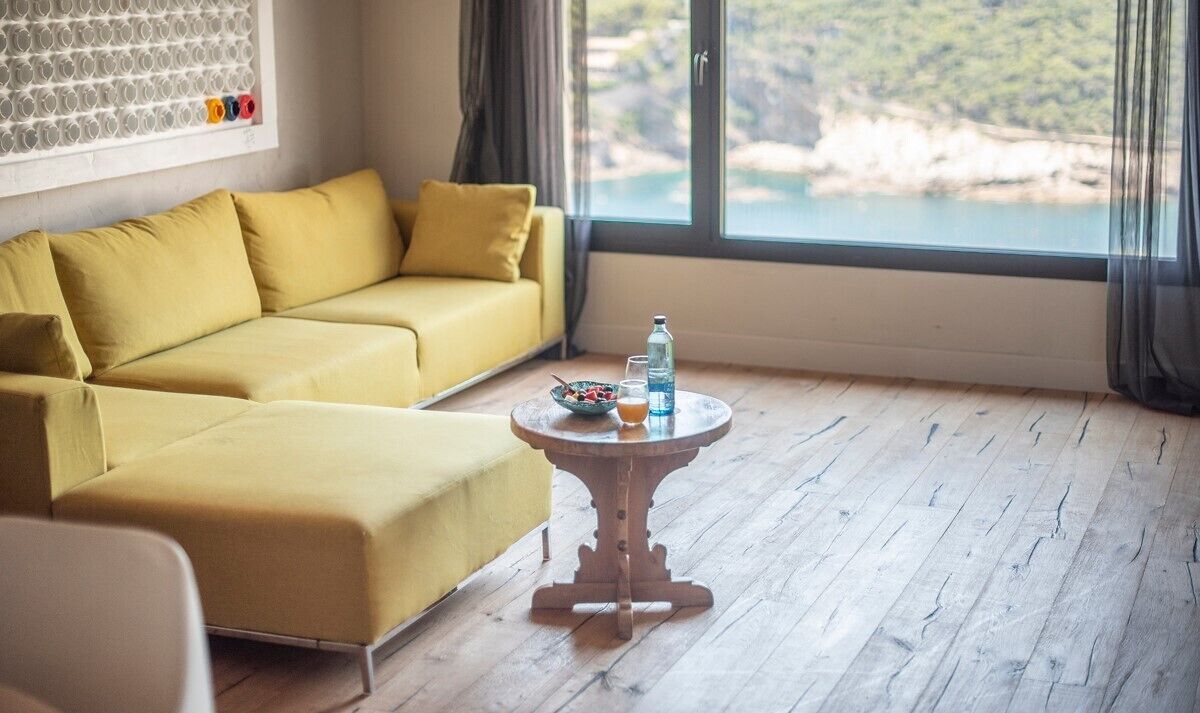 Une pièce avec un canapé jaune, une table ronde en bois sur laquelle se trouvent une bouteille et des verres, et une vue sur un paysage côtier par de grandes fenêtres.
