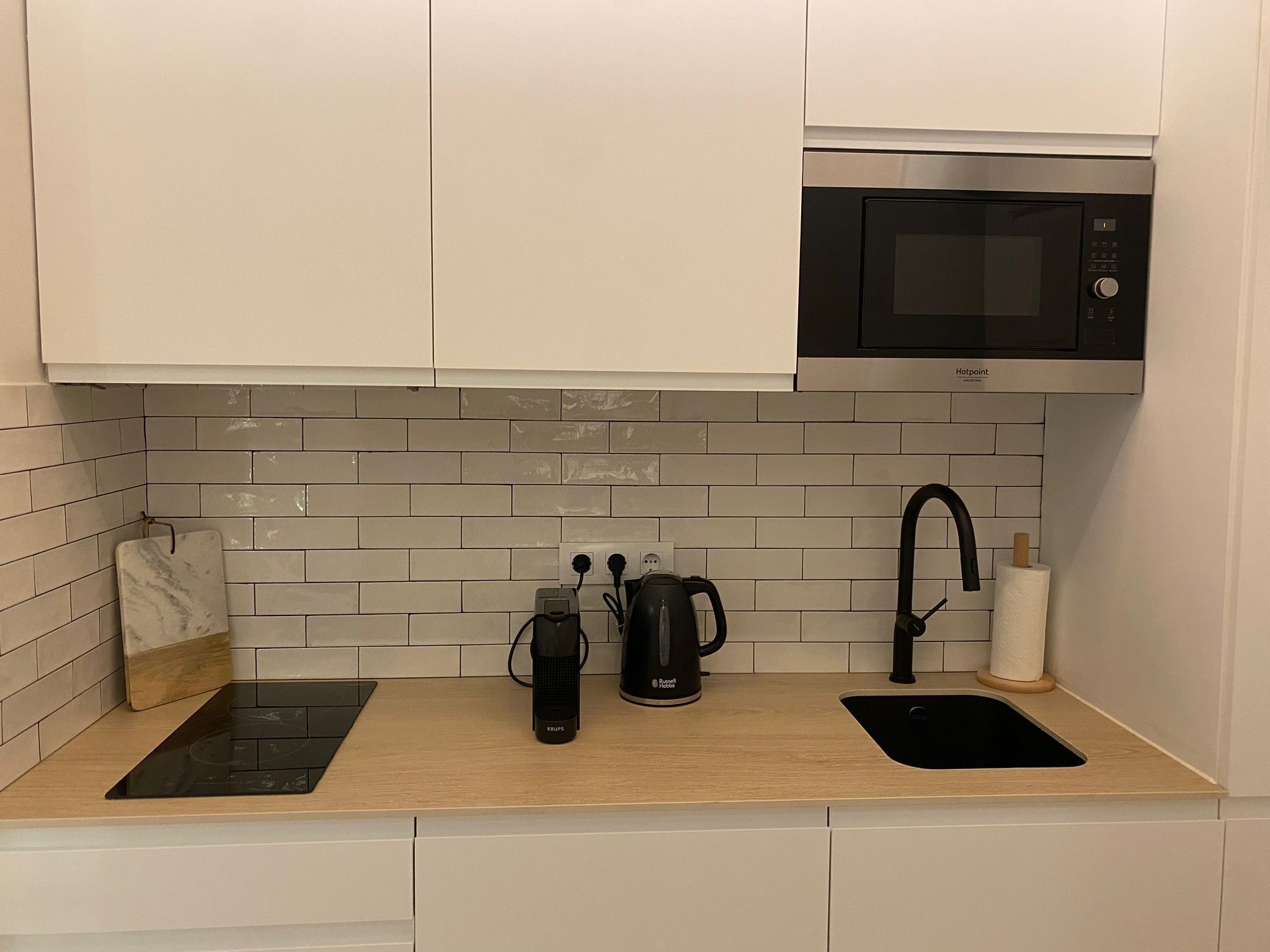 Una cocina moderna con armarios blancos, una placa de inducción negra, microondas, hervidor y pileta.