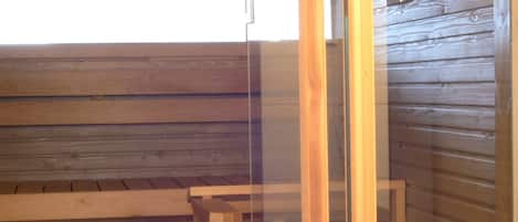 Una sala de sauna con bancos de madera, un banco con un recipiente negro y una partición de vidrio.