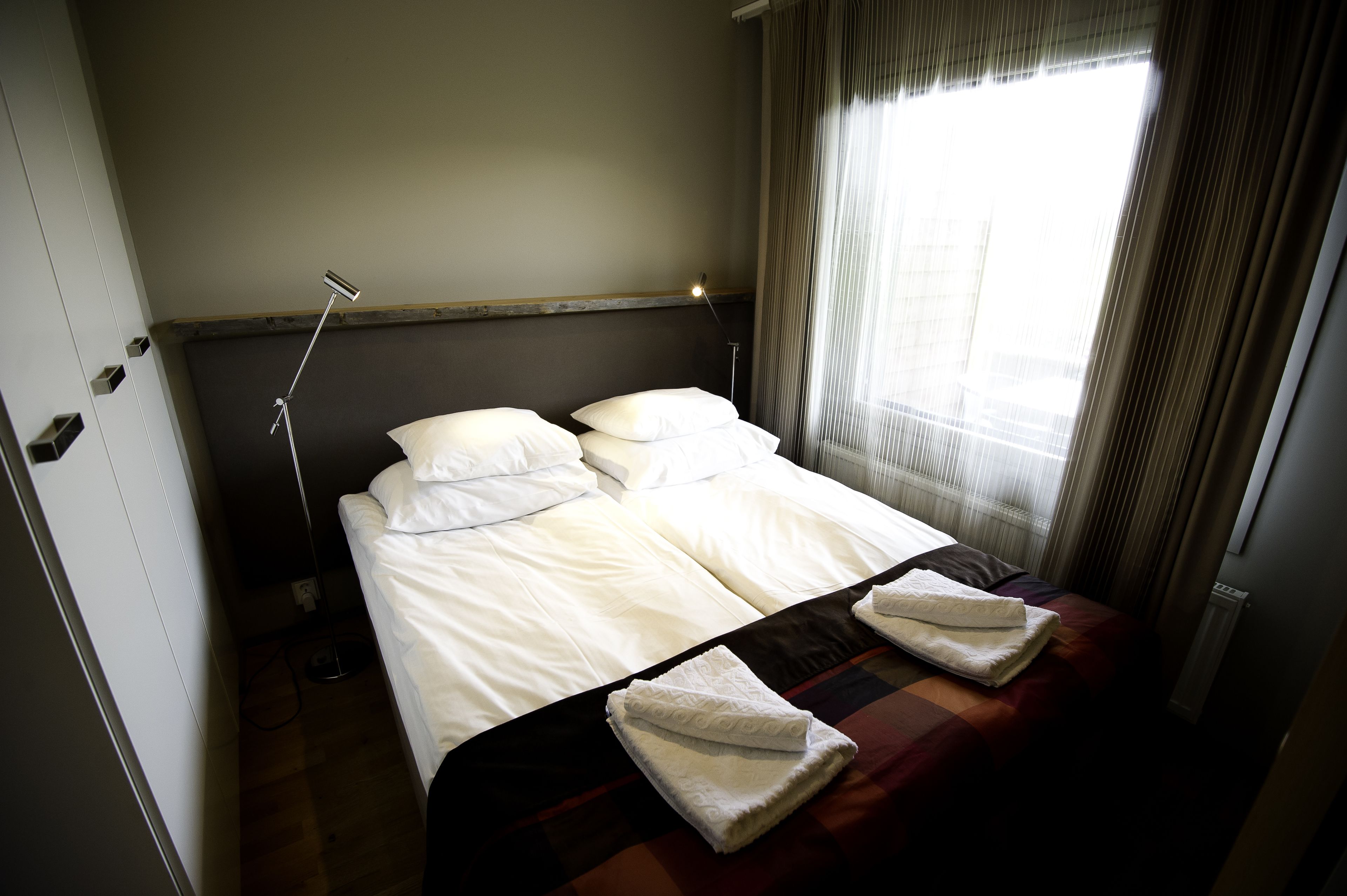A neatly made bed with white linens and two towels, a bedside lamp, and a window with curtains.