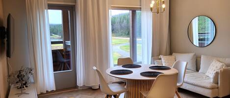 Un salón moderno con una mesa de comedor redonda, sofás blancos y un amplio ventanal con vistas a una zona boscosa.