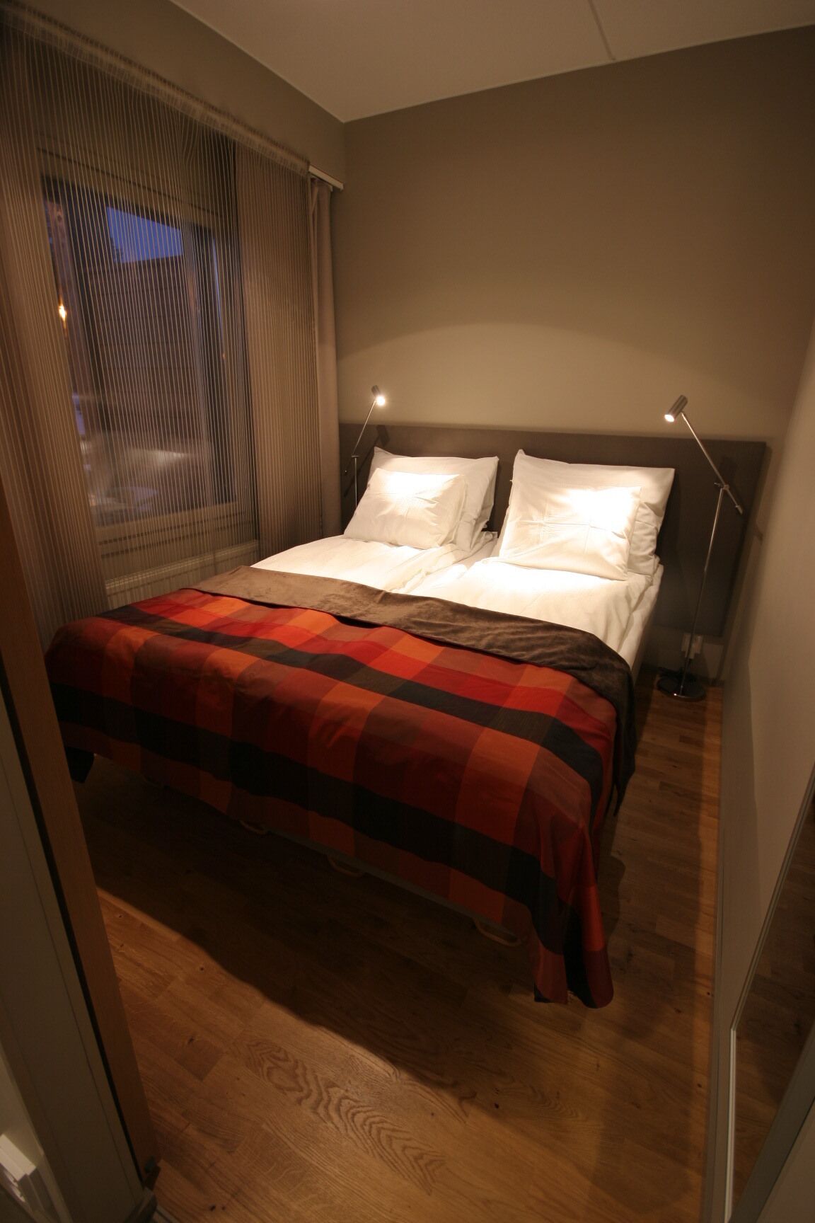 A bedroom with a bed, two bedside lamps, a window with curtains, and a wooden floor.