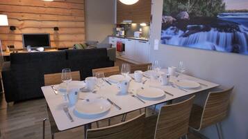 Un comedor moderno con una mesa blanca dispuesta para seis personas, rodeada de sillas de mimbre. Se ve una cocina con una pared de madera y una mesa de comedor con un cuadro de una cascada al fondo.