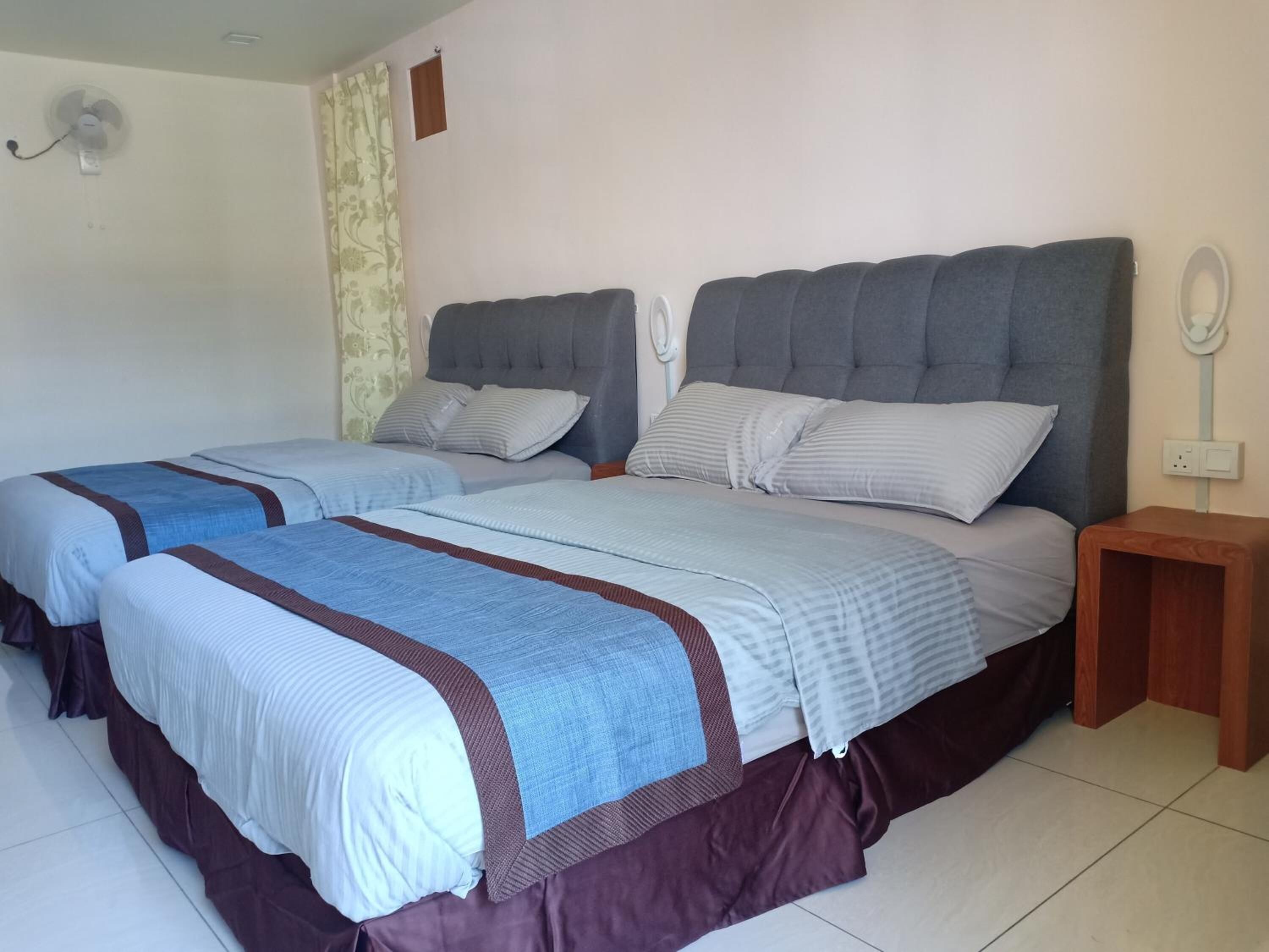 A hotel room with two beds, each with a grey headboard, white bedding, and a brown skirt. There is a small wooden nightstand with a mirror and a wall-mounted fan.